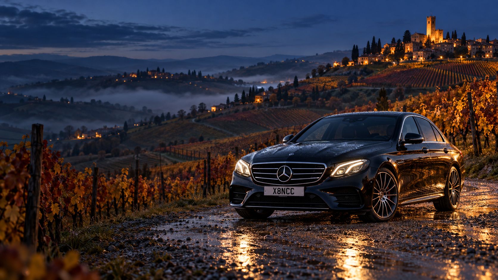 Mercedes Classe E nera su una strada sterrata tra i vigneti delle Langhe, al crepuscolo in autunno, con un borgo con torre illuminata sulla collina