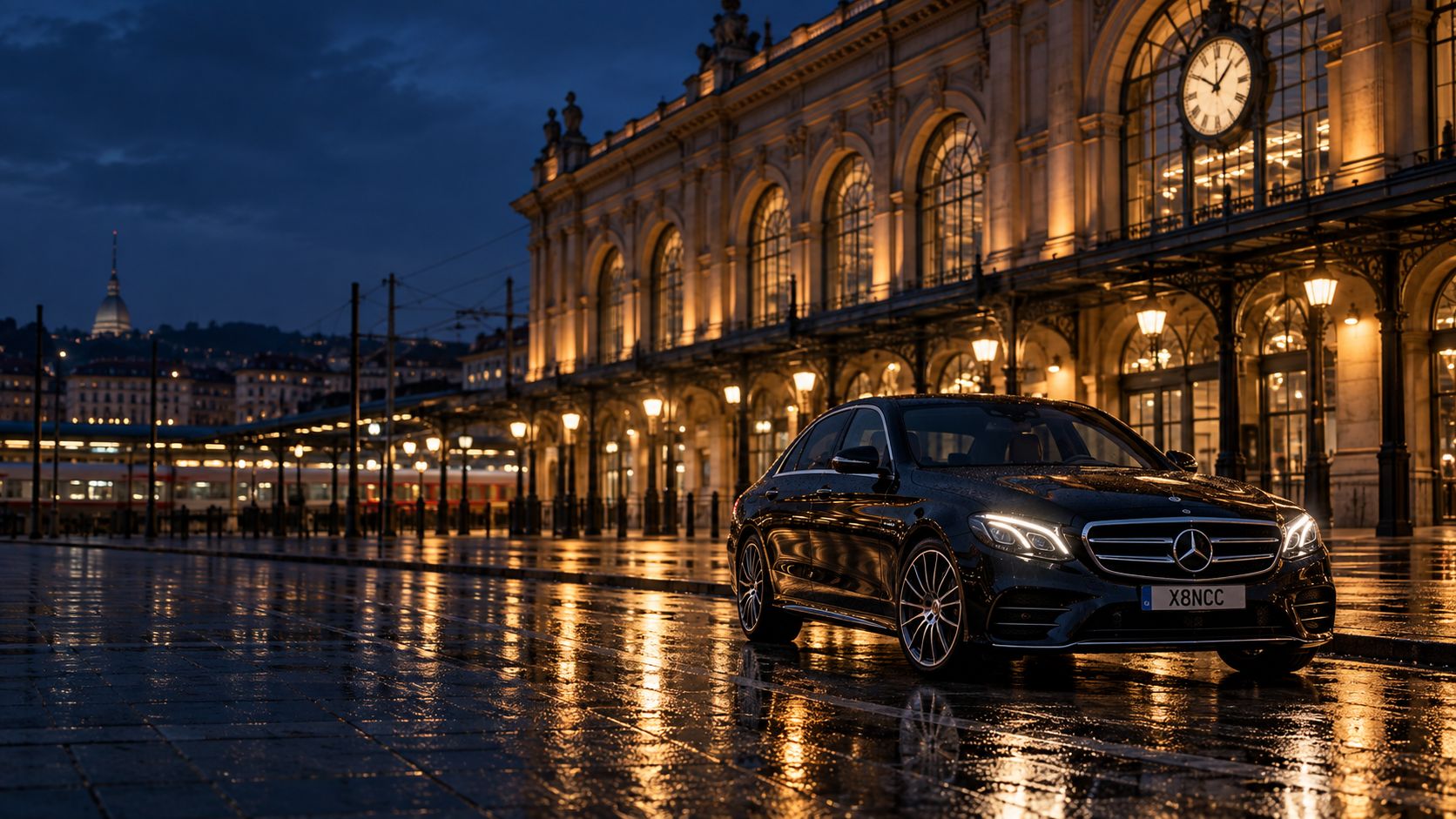 Mercedes Classe E nera ferma davanti a una grande stazione ferroviaria ottocentesca, di notte, con le grandi finestre ad arco illuminate